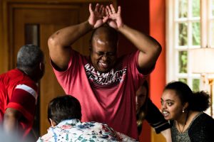 A Black man in a red t-shirt raises his hands over his head and grins