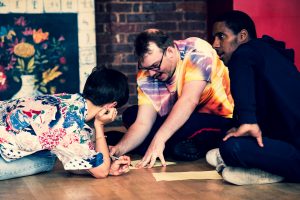Men sit on the floor drawing. Man in front wears a bright tie dye tshirt