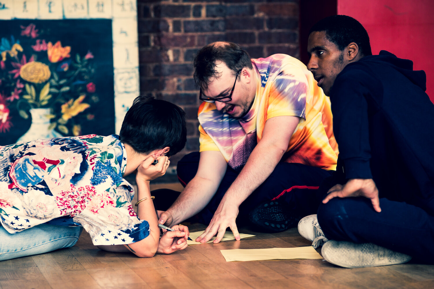 Men sit on the floor drawing. Man in front wears a bright tie dye tshirt