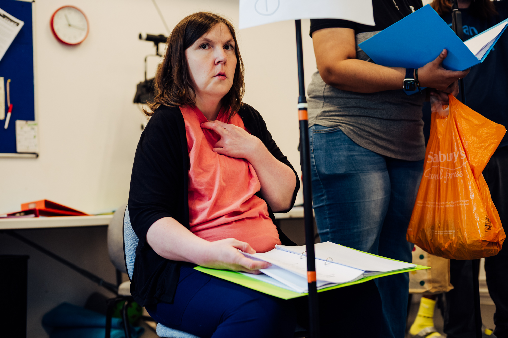 A white woman with dark brown shoulder-length hair is sitting in a bright space looking afar. She has an open script on her knees, holding the pages with one hand. Her other hand is placed on her heart.